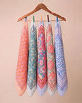 Five patterned cotton kitchen towels hanging from a wooden hanger with colorful clothespins against a solid beige background.