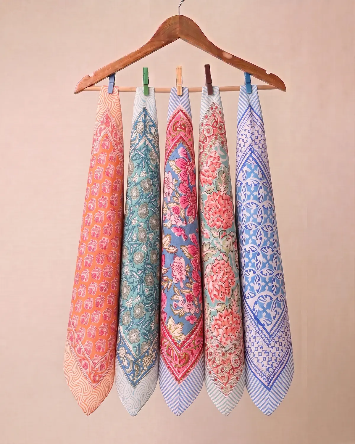 Five patterned cotton kitchen towels hanging from a wooden hanger with colorful clothespins against a solid beige background.
