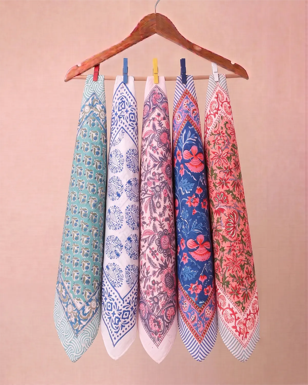 Five patterned square cotton scarves in various colors (teal, white, pink, blue, red) hanging from a wooden hanger on a solid beige background.