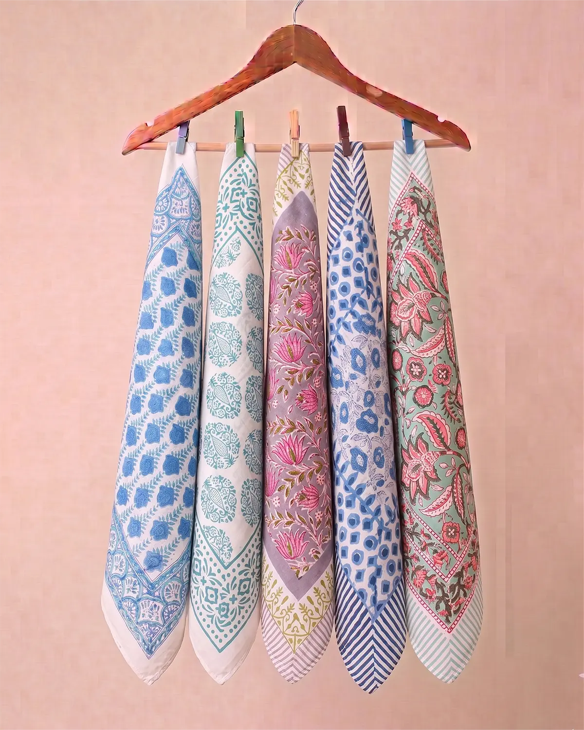 Five patterned cotton hand block printed bandana in blue, white, pink, and purple hanging from a wooden hanger against a soft pink background.