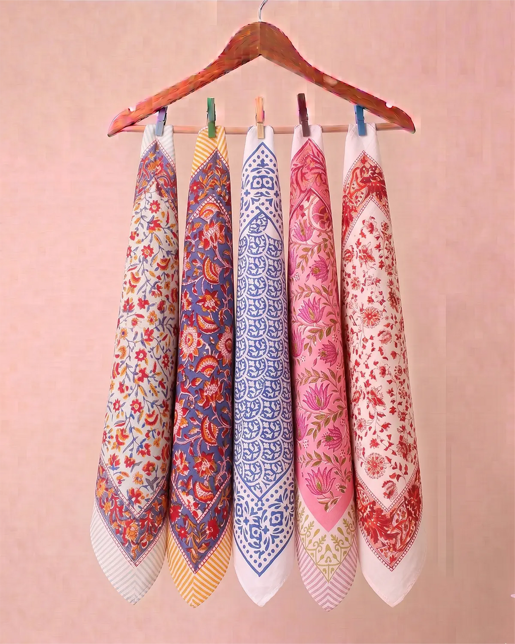 Five patterned hand block printed bandana hanging from a wooden hanger against a soft pink wall. Each towel has a different floral and geometric design in colors including red, blue, pink, yellow, and white.