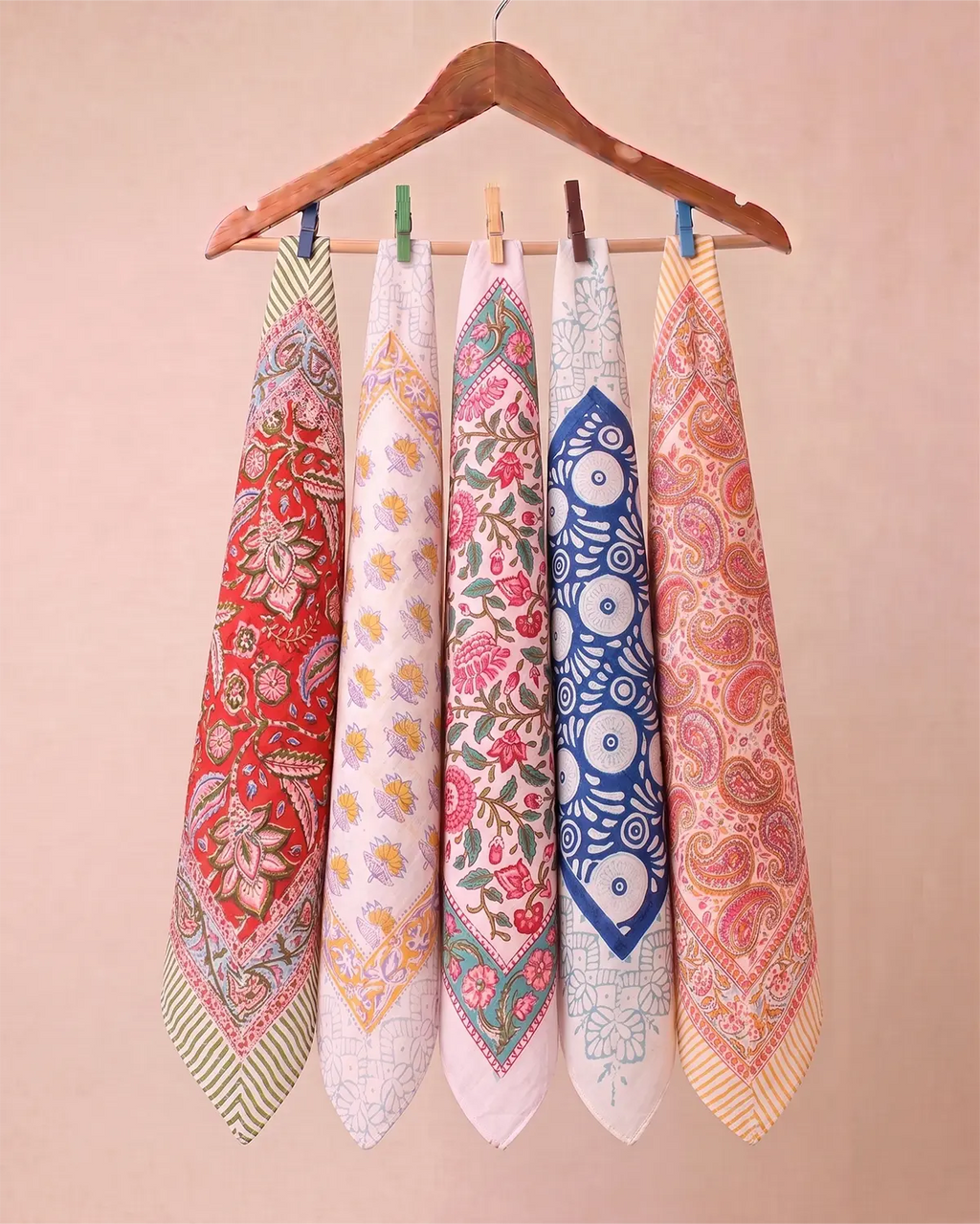 Five patterned cotton Hand block printed bandana in red, white, pink, blue, and peach hang from a wooden hanger with clothespins against a soft beige background.