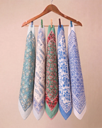Five patterned square cotton hand block printed bandana scarves or bandanas in assorted colors hang from a wooden hanger with wooden and metal clothespins against a plain beige wall.