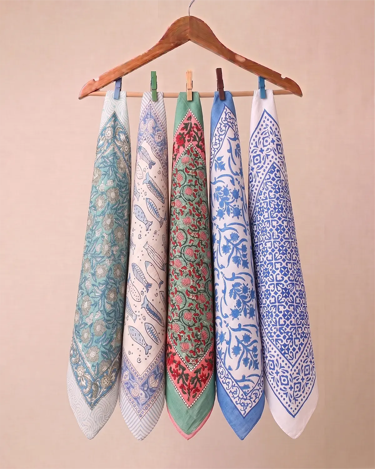 Five patterned square cotton hand block printed bandana scarves or bandanas in assorted colors hang from a wooden hanger with wooden and metal clothespins against a plain beige wall.