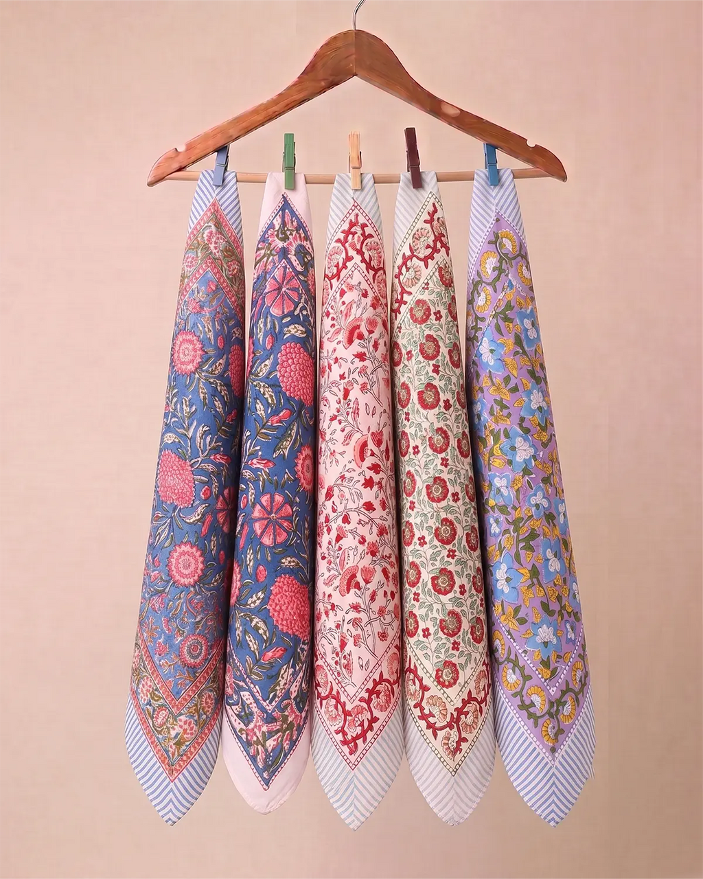 Five patterned square hand block printed bandanas with striped borders hang from a wooden hanger against a solid beige background.