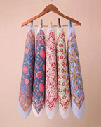Five patterned square hand block printed bandanas with striped borders hang from a wooden hanger against a solid beige background.