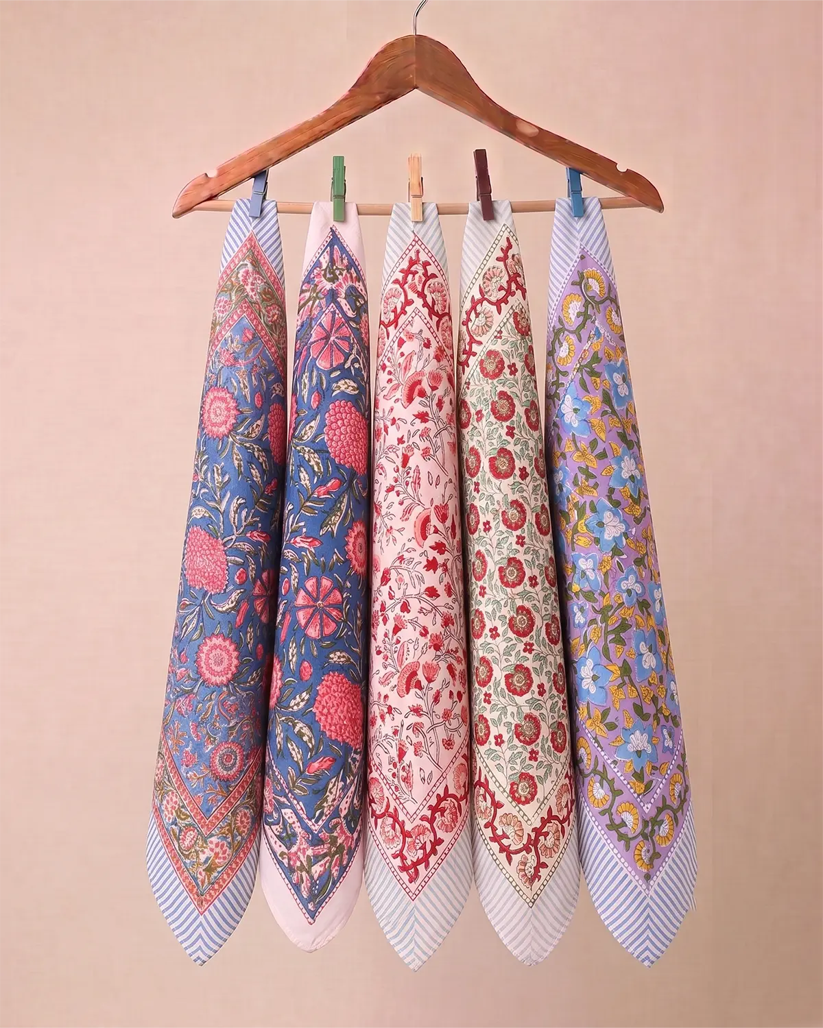 Five patterned square hand block printed bandanas with striped borders hang from a wooden hanger against a solid beige background.