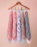Five patterned cotton kitchen towels hanging from a wooden hanger with wooden pegs against a solid beige background.
