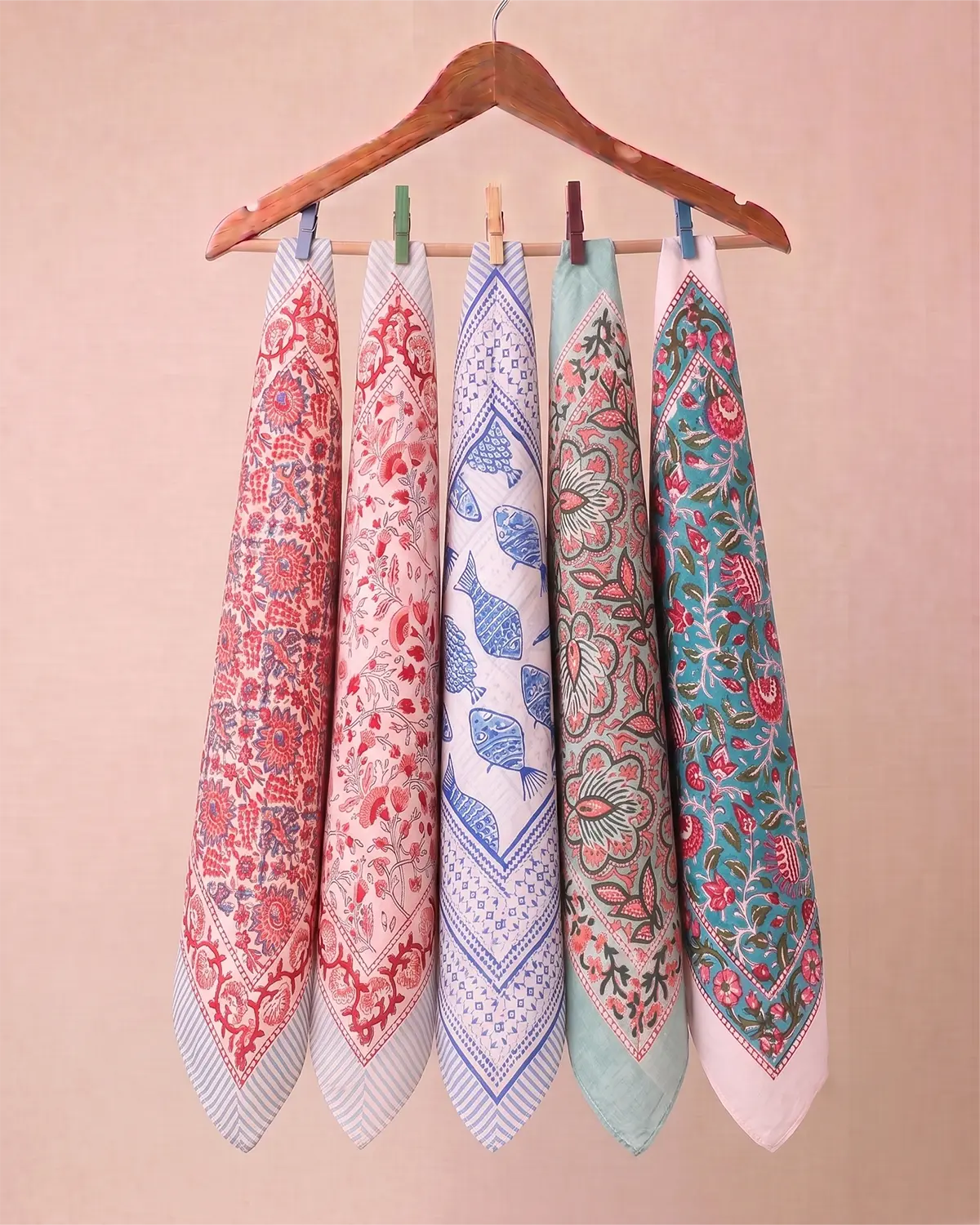 Five patterned cotton kitchen towels hanging from a wooden hanger with wooden pegs against a solid beige background.