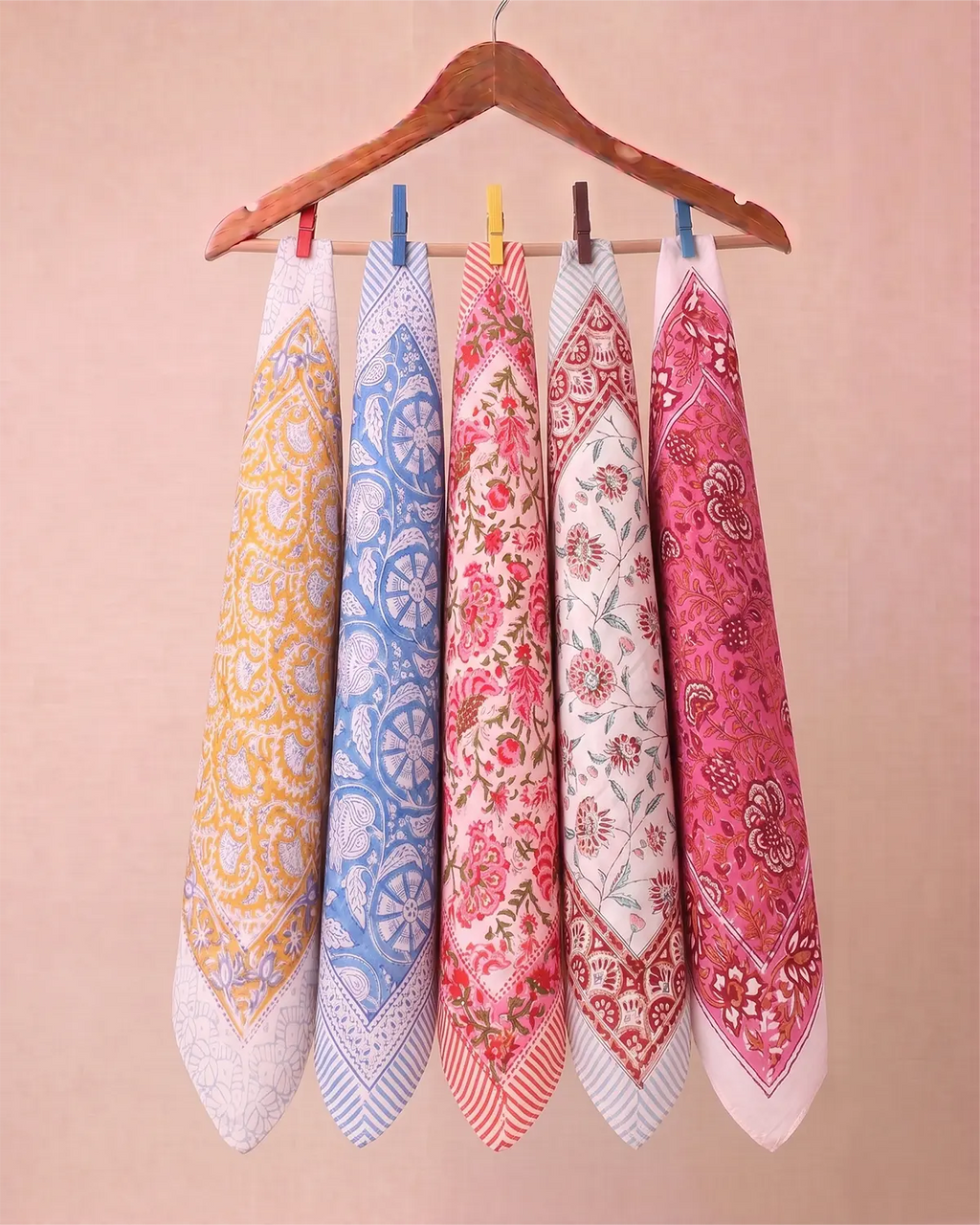 Five patterned cotton hand block printed bandana in yellow, blue, red, and pink hanging from a wooden hanger against a solid beige background.
