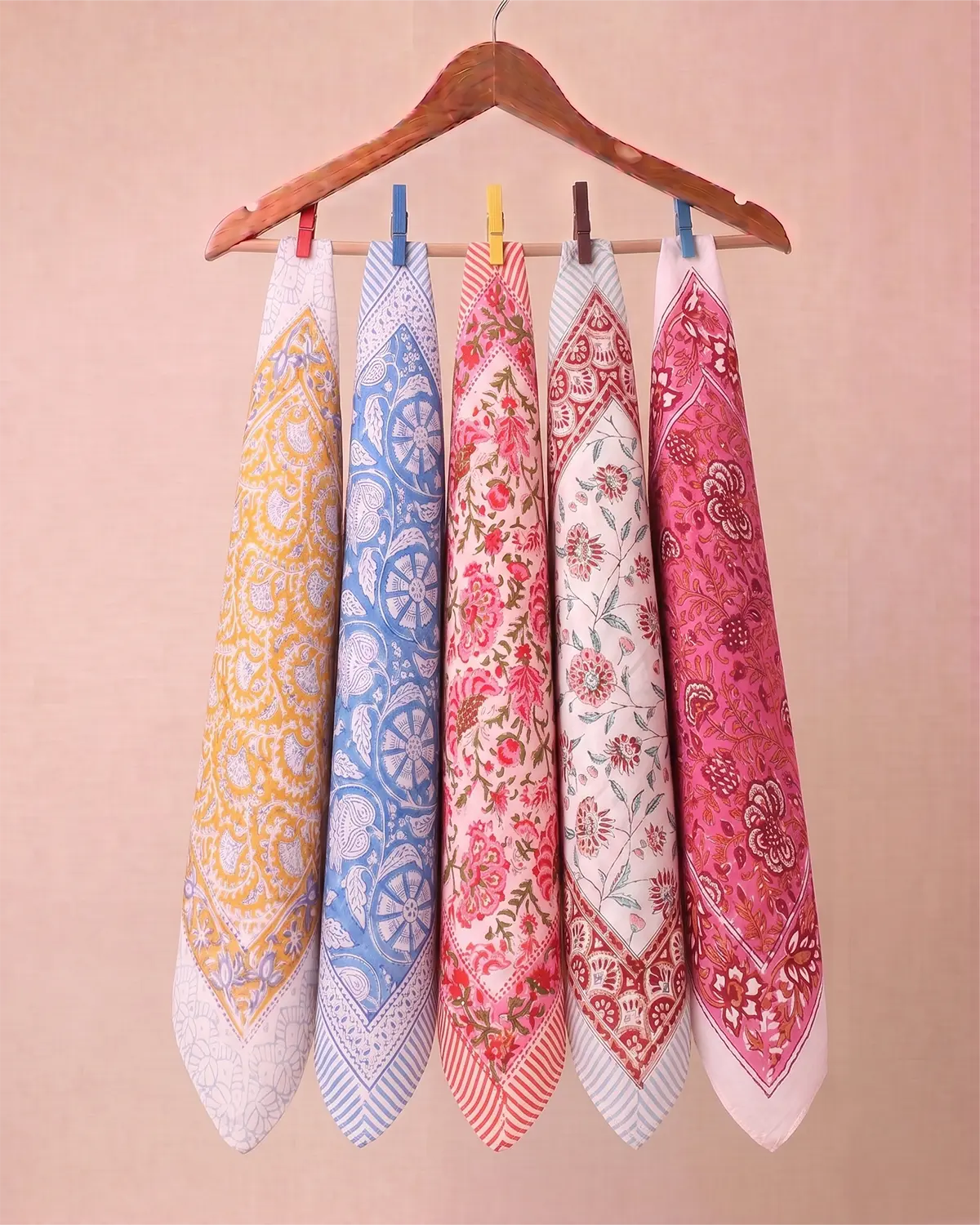 Five patterned cotton hand block printed bandana in yellow, blue, red, and pink hanging from a wooden hanger against a solid beige background.